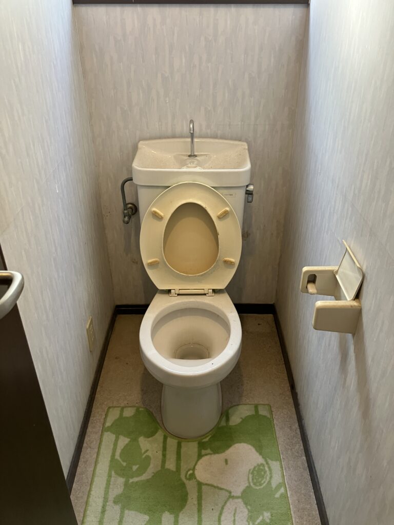 2階トイレの施工前。便器・手洗いタンク・壁紙に経年の汚れが目立ち、床のクッションフロアも色あせています。